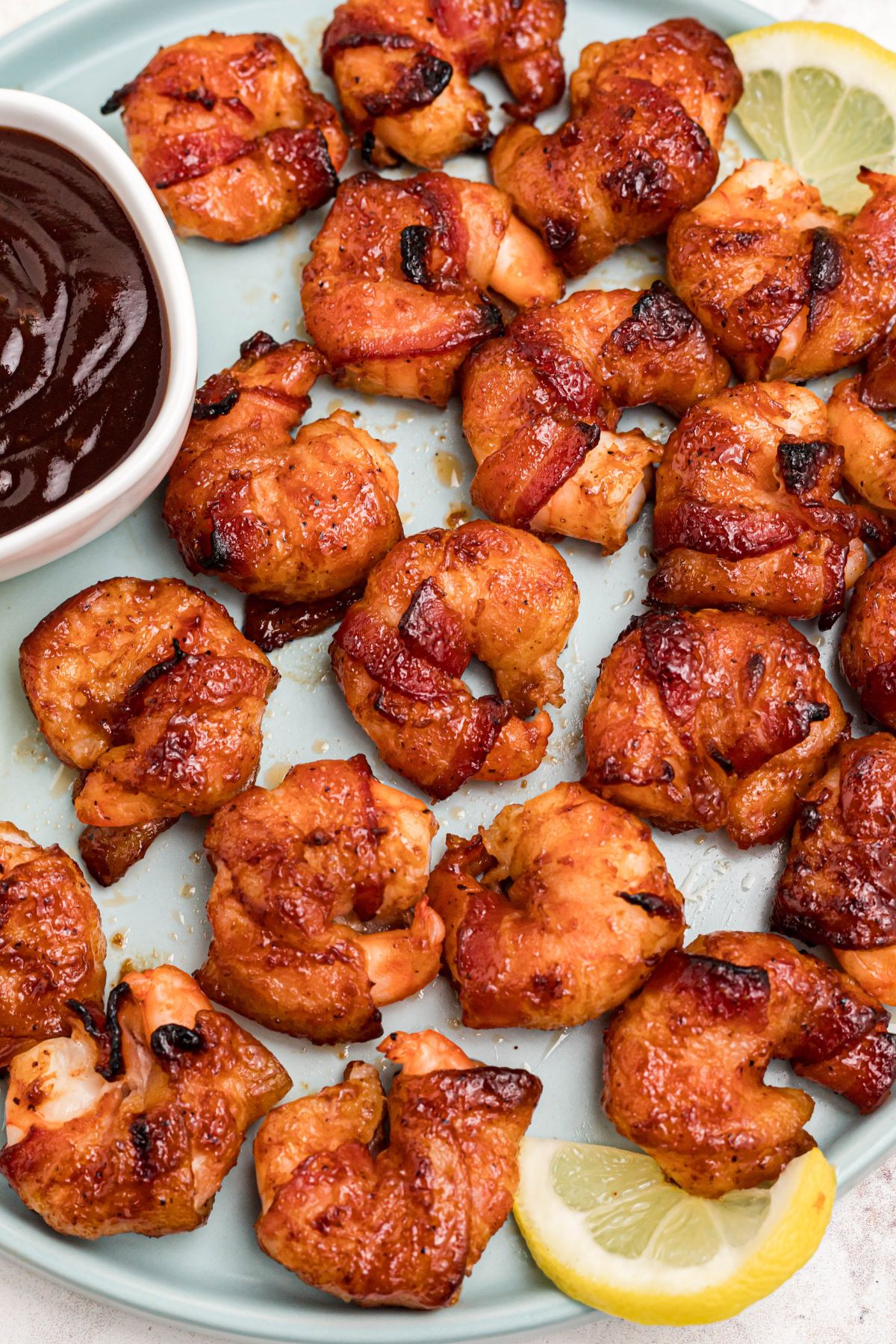 Crispy bacon wrapped around shrimp, coated with BBQ sauce mixture, on a light green plate with lemon slices and serving tongs on the table.