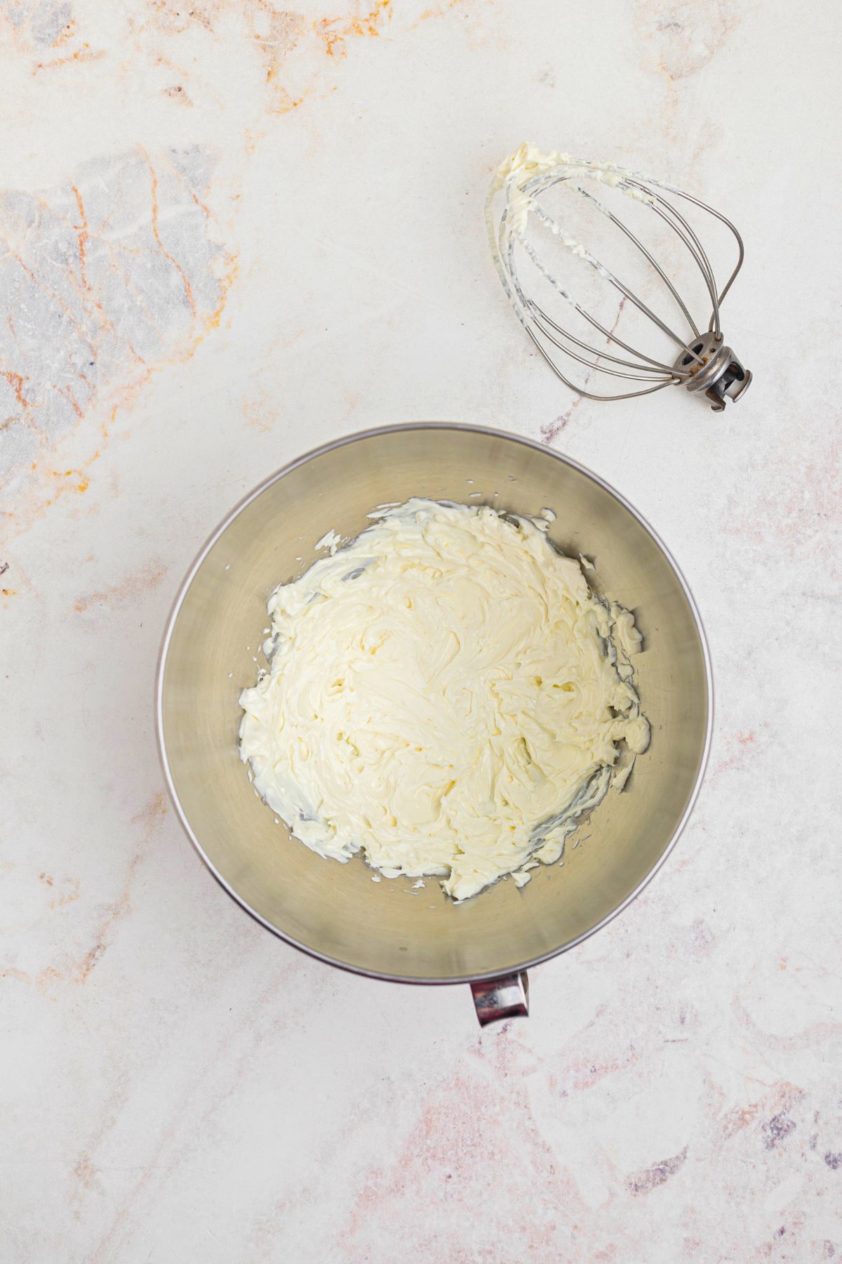 Cream cheese beaten in a silver mixing bowl, until smooth and creamy, on a white marble table. 