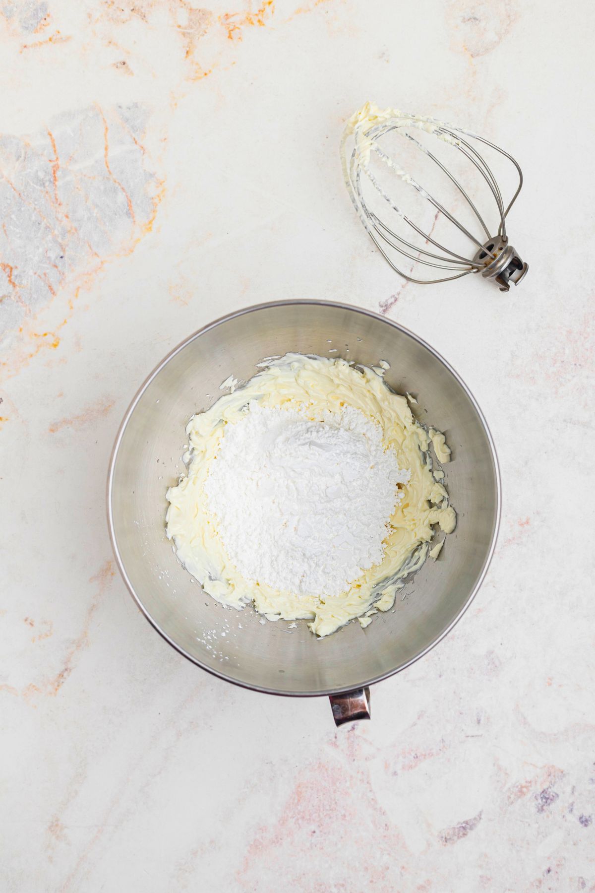 Powdered sugar being added to cream cheese in a silver mixing bowl. 