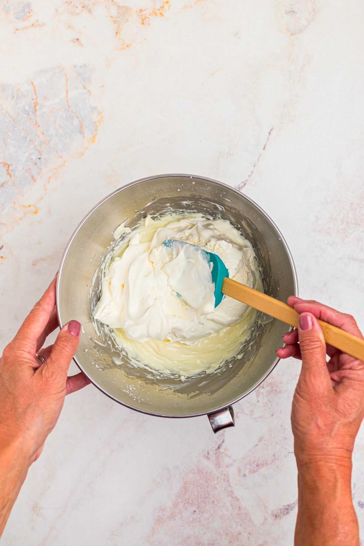 Cool whip being mixed in to cream cheese mixture in a silver mixing bowl, with a spatula. 