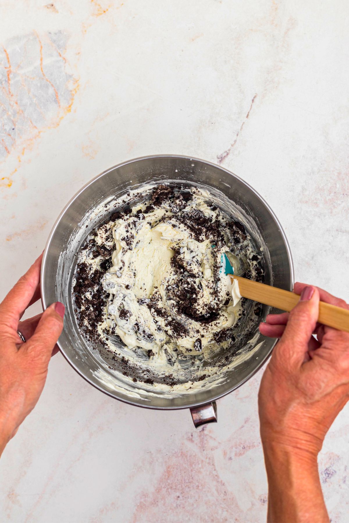 Crushed Oreos being stirred into cream cheese mixture in a silver mixing bowl. 