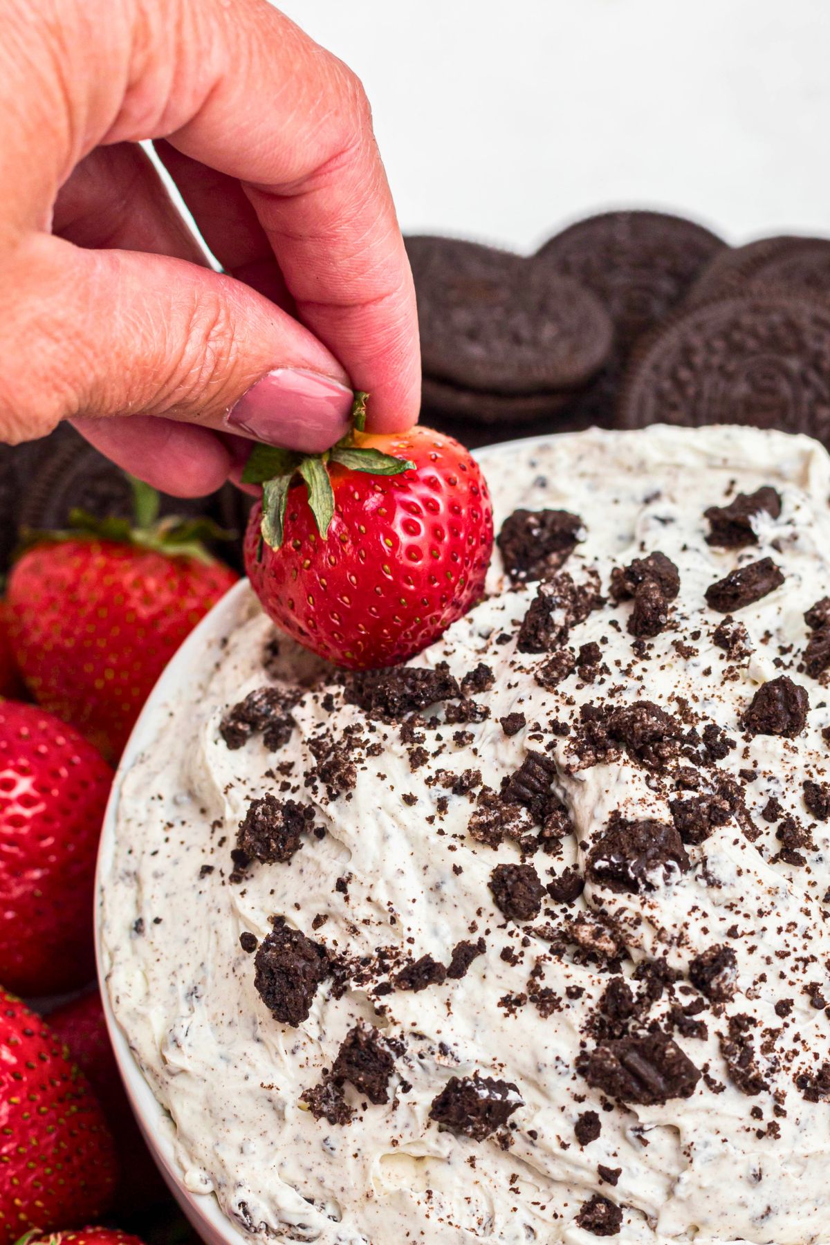 A hand dipping a strawberry into Oreo dip, surrounded by more strawberries, Oreos, and pretzels. 
