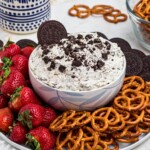 Strawberries, pretzels, and chocolate cookies, on a round white plate, around a cookies and cream Oreo dip, on a marble table.