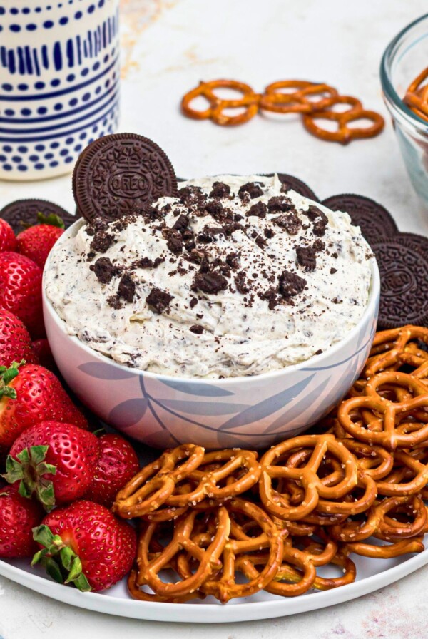 Strawberries, pretzels, and chocolate cookies, on a round white plate, around a cookies and cream Oreo dip, on a marble table.
