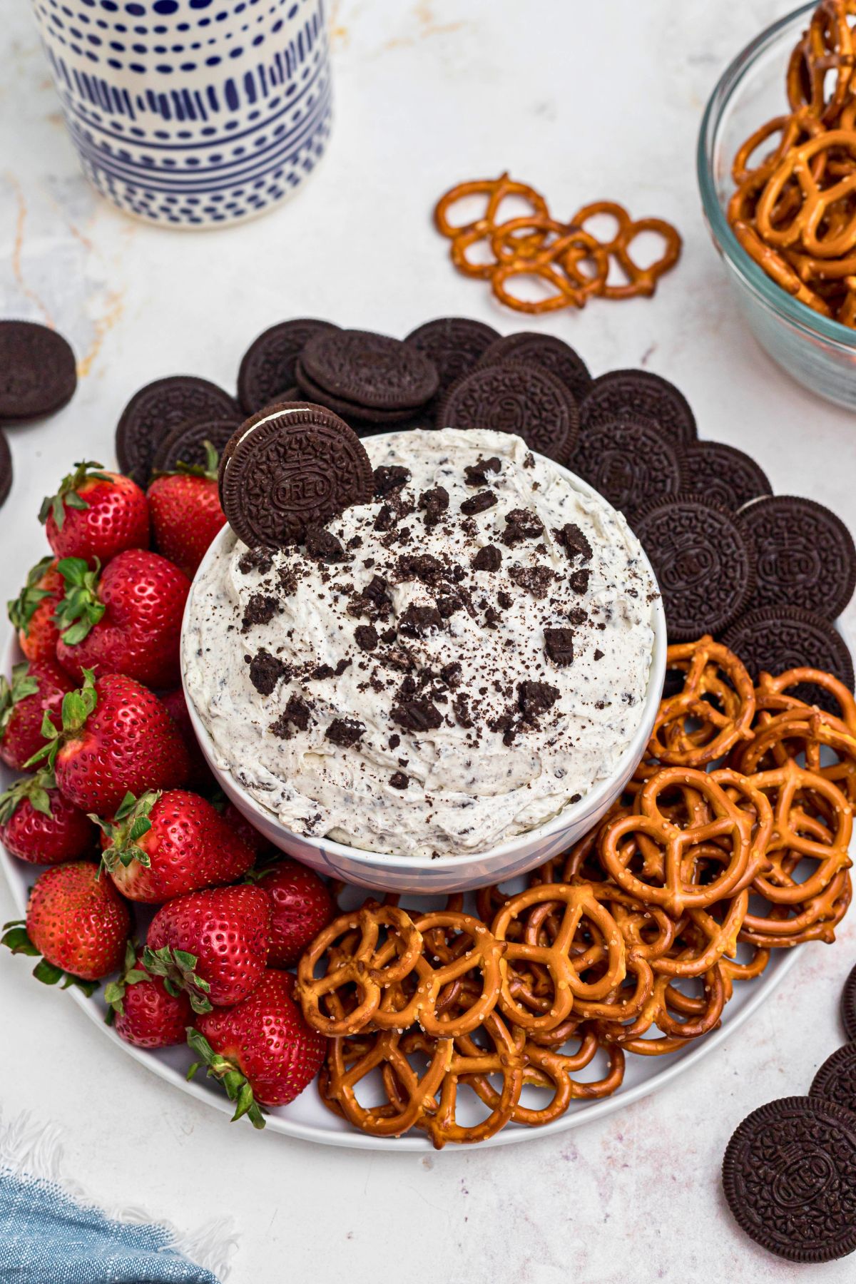 Strawberries, pretzels, and chocolate cookies, on a round white plate, around a cookies and cream Oreo dip, on a marble table. 