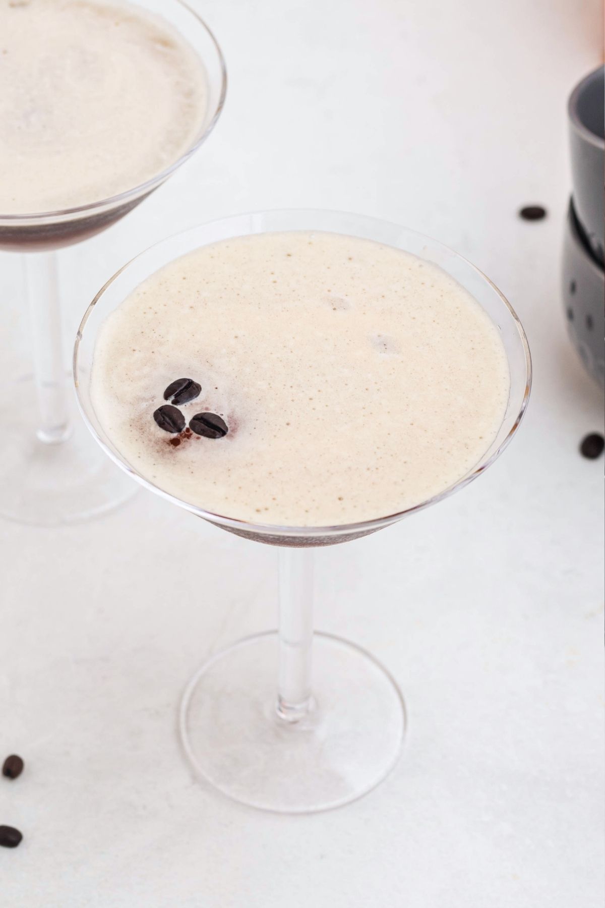 Froth on top of an espresso and vodka mixture, in a martini glass, with coffee beans spread around the table, and three coffee beans on top of the foam. 