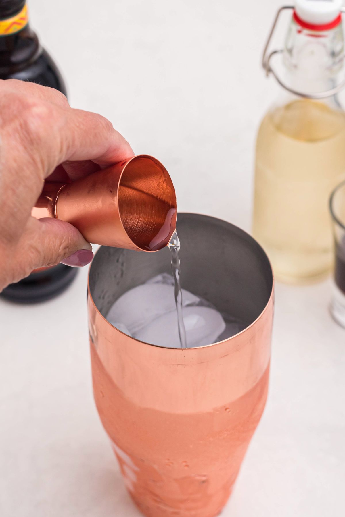 Vodka, measured and poured into a brass cocktail shaker over ice, on a white marble table. 