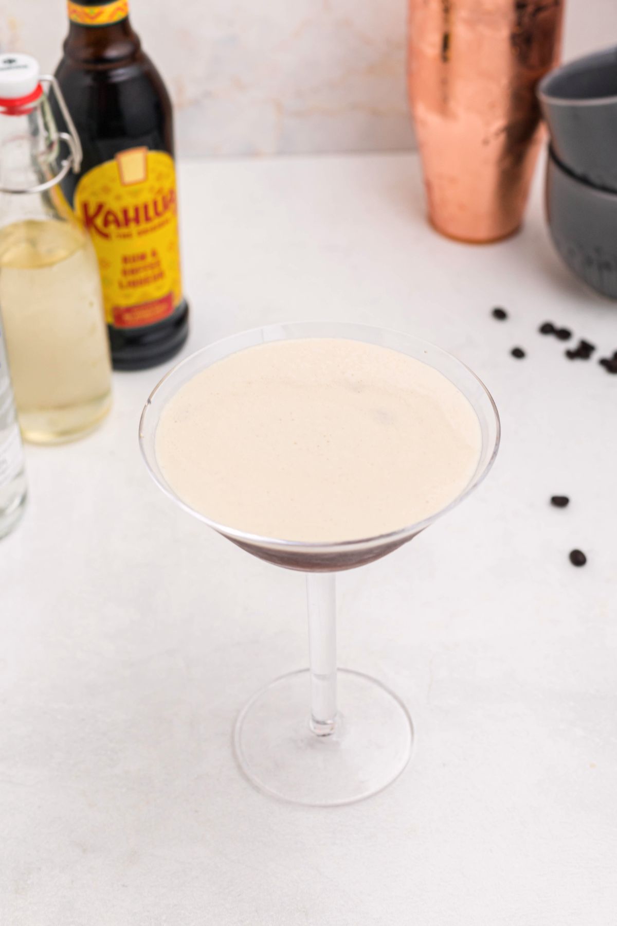 Froth on top of an espresso and vodka mixture, in a martini glass, with coffee beans spread around the table. 