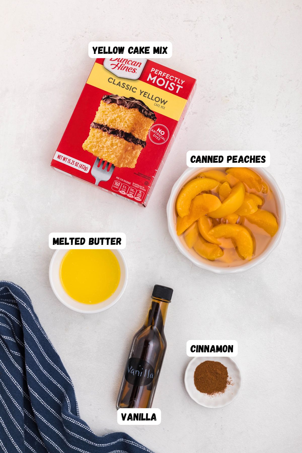 Boxed cake mix, peaches, butter, vanilla, and cinnamon, in small bowls on a white marble table. 