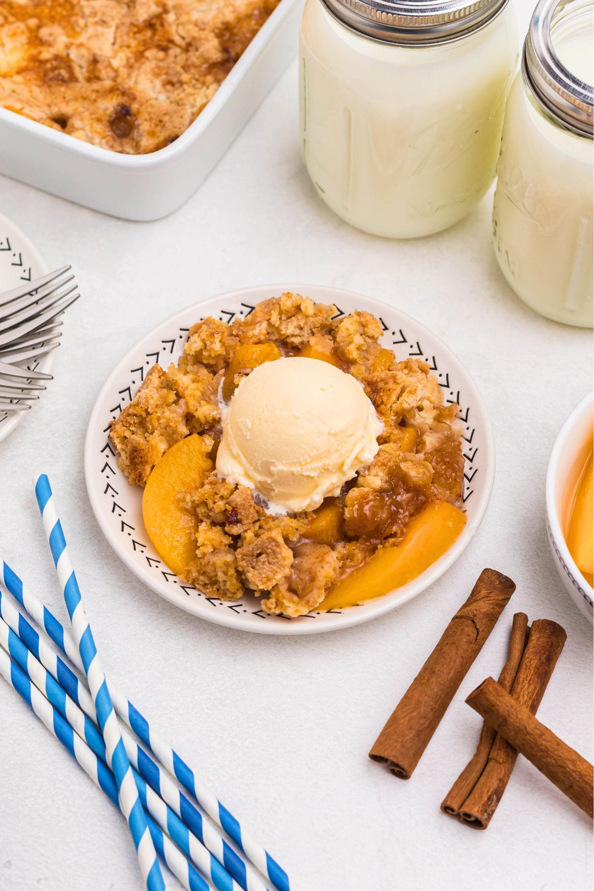 Peach cobbler scooped out and served on a small plate, in front of the whole dish, with blue striped straws and cinnamon sticks on the table.