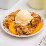 Peach cobbler scooped out and served on a small plate, in front of the whole dish, with blue striped straws and cinnamon sticks on the table.