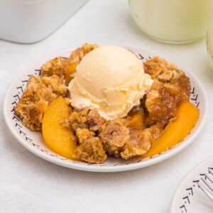 Peach cobbler scooped out and served on a small plate, in front of the whole dish, with blue striped straws and cinnamon sticks on the table.
