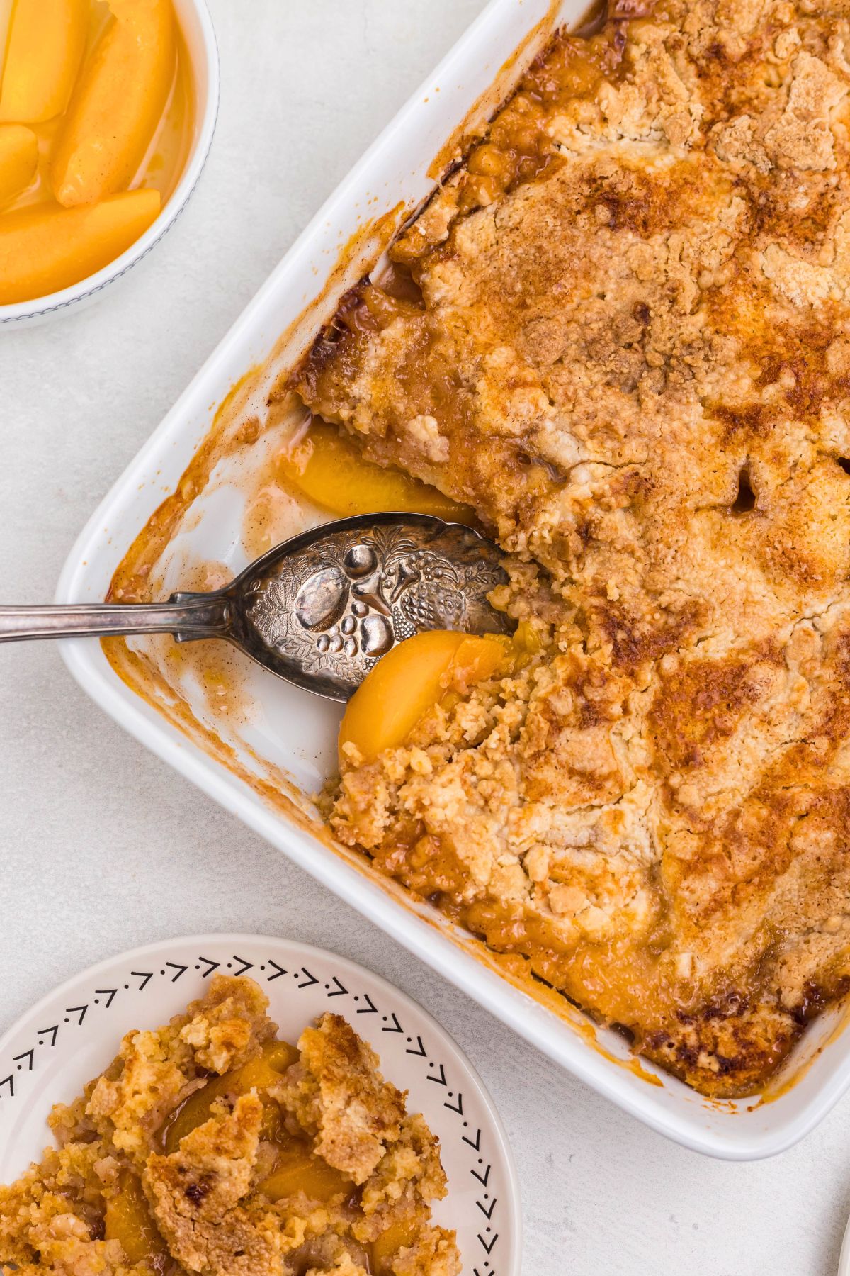 Peach dump cake with a slice spooned out onto s small white plate, with the serving spoon in the dish. 
