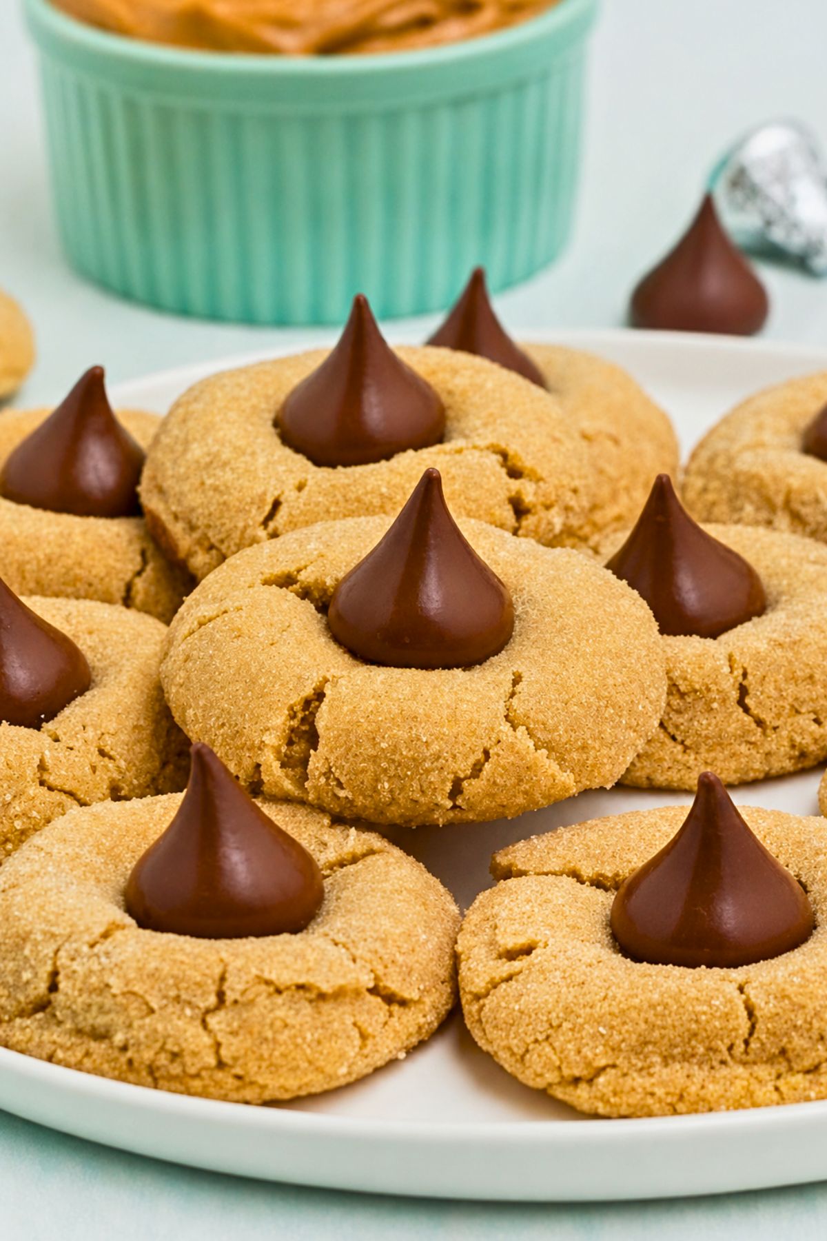 Peanut Butter cookies, topped with a chocolate kiss, stacked on a white plate, with a bowl of peanut butter on the table.