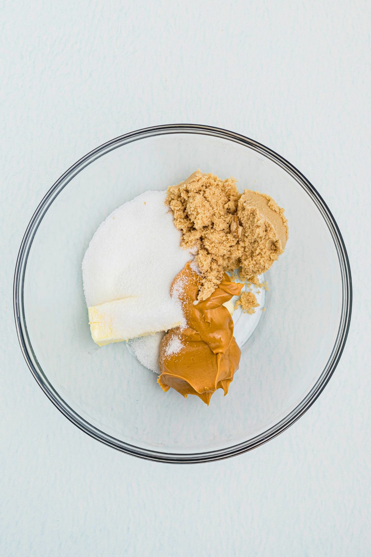 Butter, sugar, peanut butter, and brown sugar, in a large clear mixing bowl, before being mixed to make cookies. 