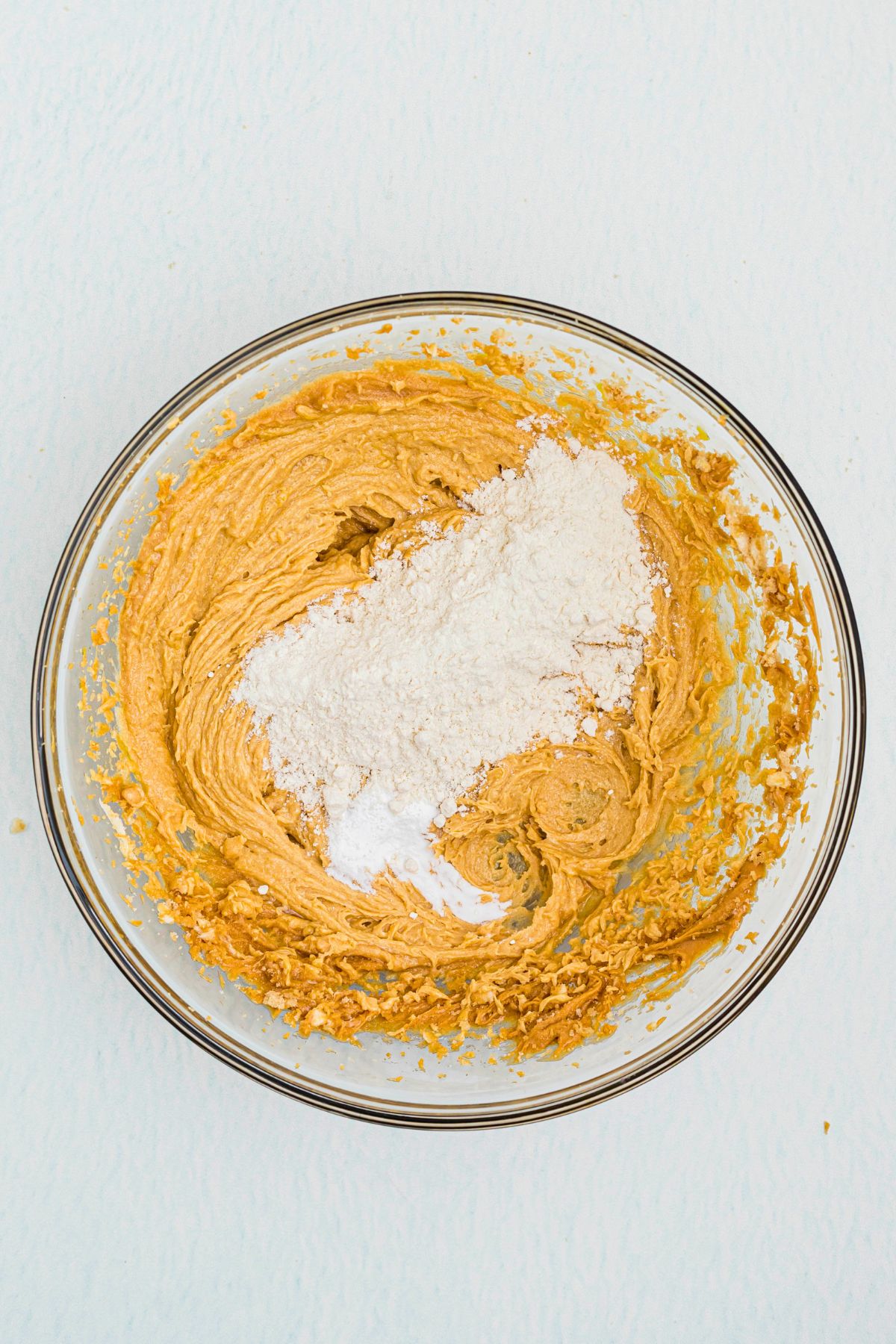 Flour, baking powder, and salt being added to peanut butter cookie dough in a clear glass bowl. 