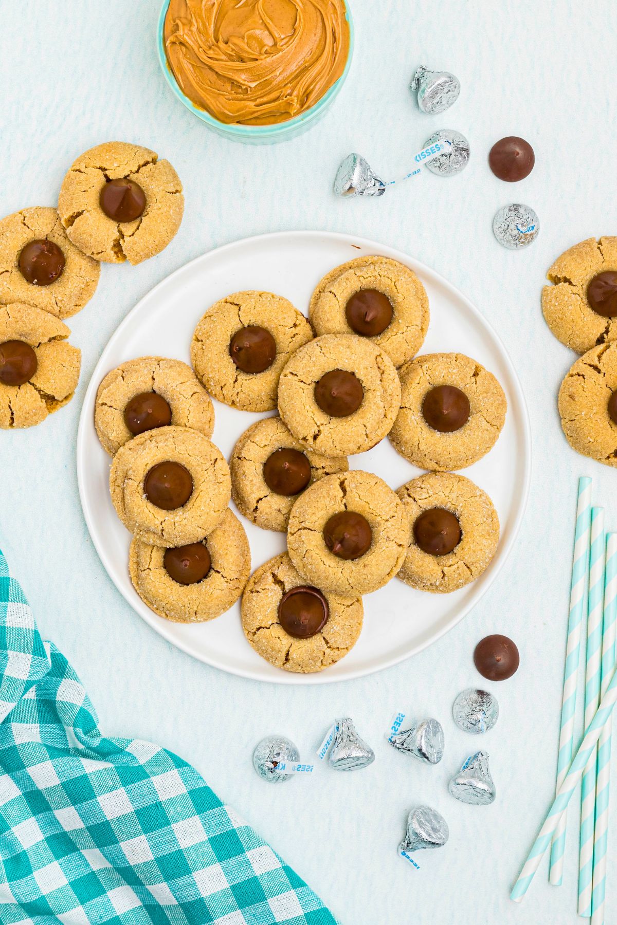 Peanut Butter cookies, topped with a chocolate kiss, stacked on a white plate, with a bowl of peanut butter on the table, next to a teal checked linen. 