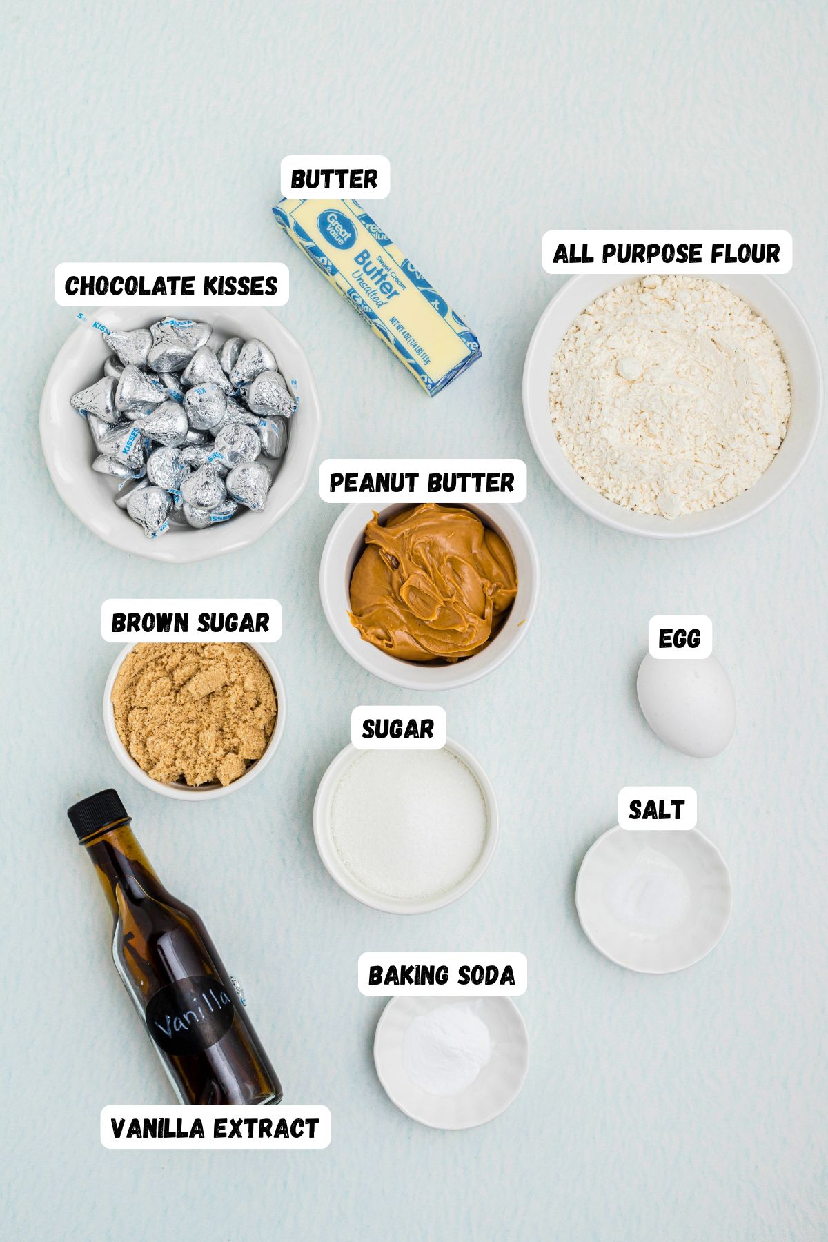 Creamy peanut butter, Unsalted butter, Granulated sugar, Light Brown sugar, Egg, Vanilla extract, All-purpose flour, Baking soda, Salt, and Hershey's, Kisses in small bowls, on a light blue table, with all ingredients labeled.