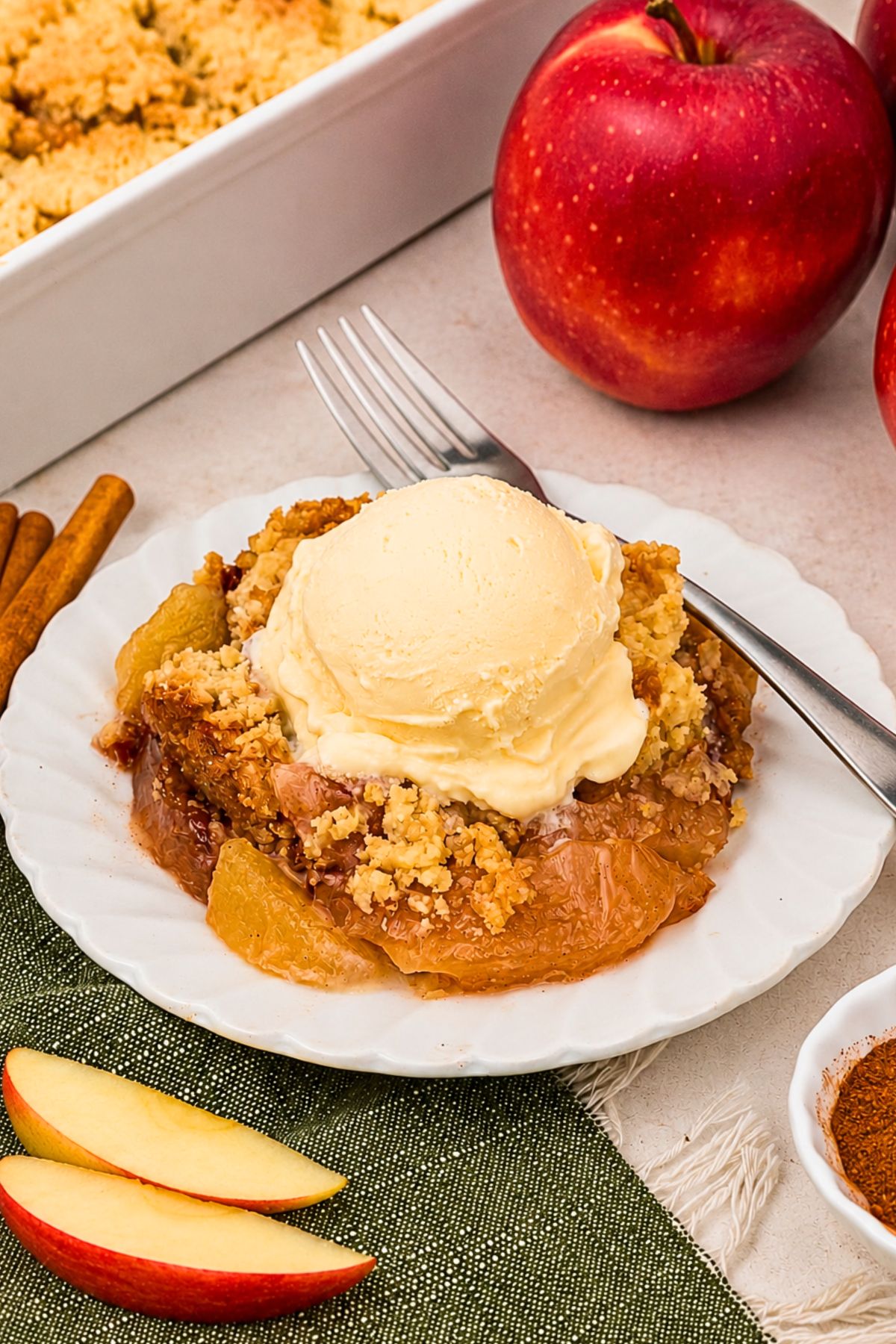 Slice of apple dump cake, topped with a scoop of ice cream, on a small round white plate, with apple slices in front of the plate.