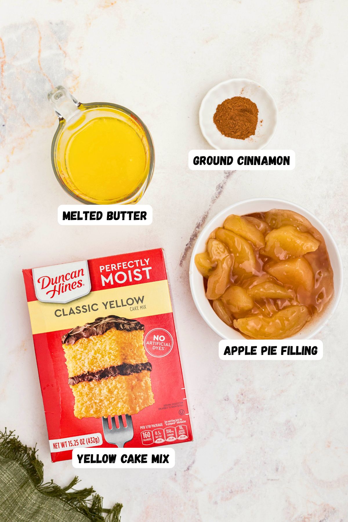 Box cake mix, melted butter, cinnamon, and apple pie filling, on a marble table, labeled, with a green linen on the table. 
