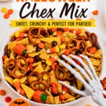 Large white bowl filled with Chex mix, pretzels, candy corn, and candies, with some scattered around the table and a scooper skeleton hand on top of the bowl.
