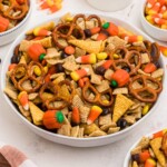 Large white bowl filled with Chex mix, pretzels, candy corn, and candies, with some scattered around the table.