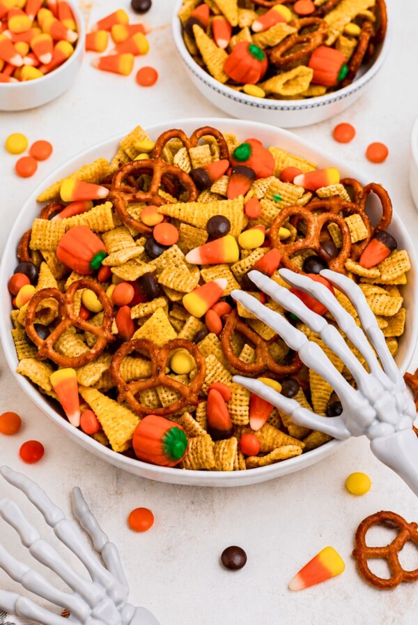 Large white bowl filled with Chex mix, pretzels, candy corn, and candies, with some scattered around the table and a scooper skeleton hand on top of the bowl.
