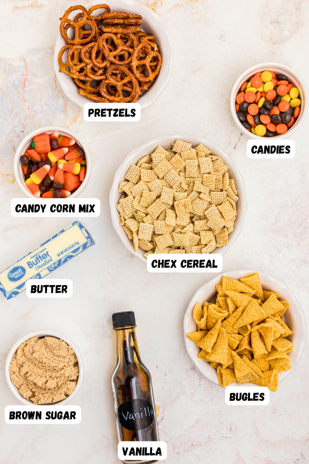 Candy corn, chex mix, bugles, pretzels, brown sugar, butter, and vanilla, in small bowls on a marble table. 