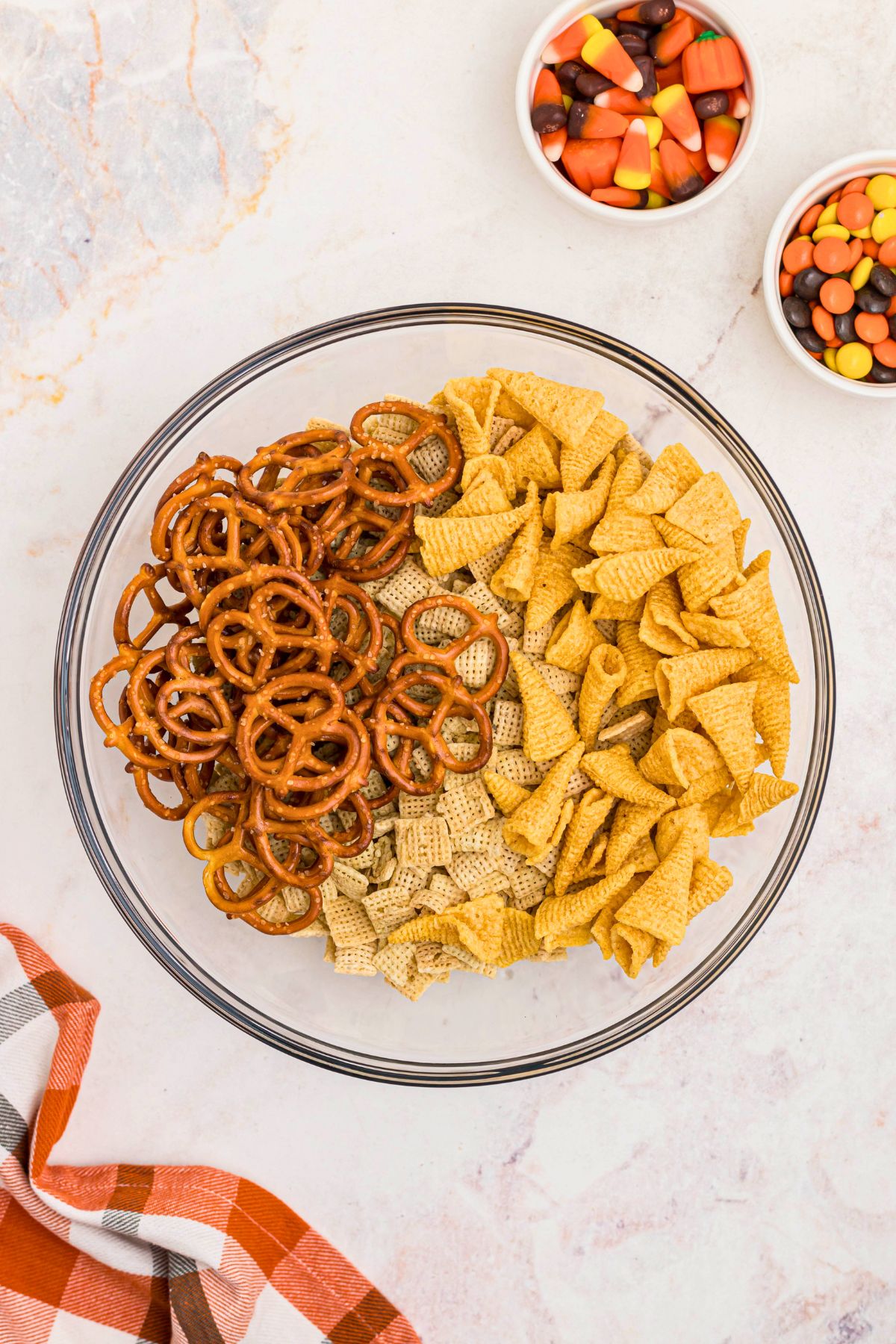 Cereal, pretzel, and bugles, in a large clear mixing bowl, with candies in small white bowls on the table. 