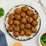 Black plate topped with white rite and juicy meatballs surrounded by chopped onions and sesame seeds on a marble table.