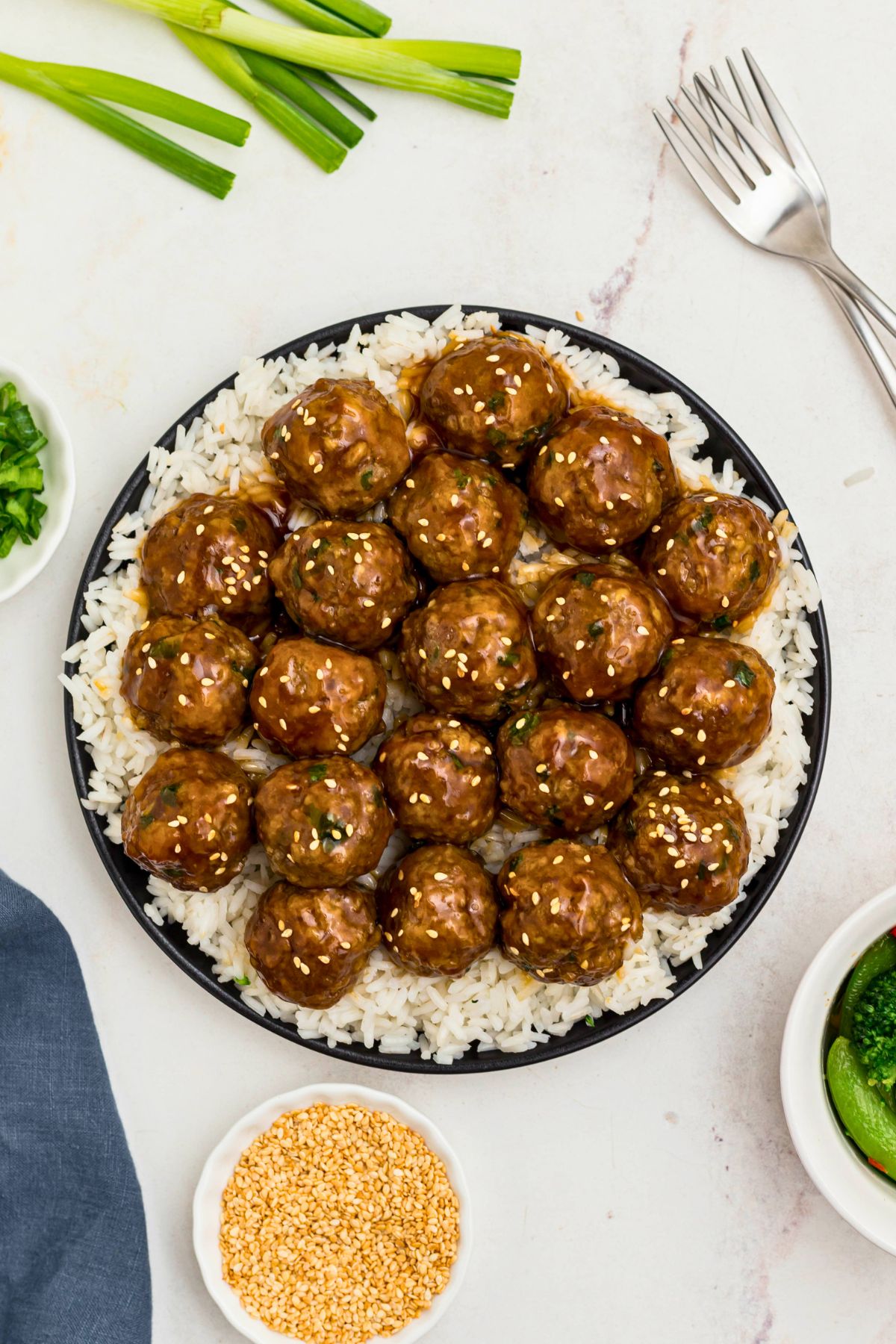 Black plate topped with white rite and juicy meatballs surrounded by chopped onions and sesame seeds on a marble table.