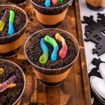 Small individual cups filled with crushed cookies, whipped cream, and chocolate pudding, topped with gummy worms, on a wooden cutting board.