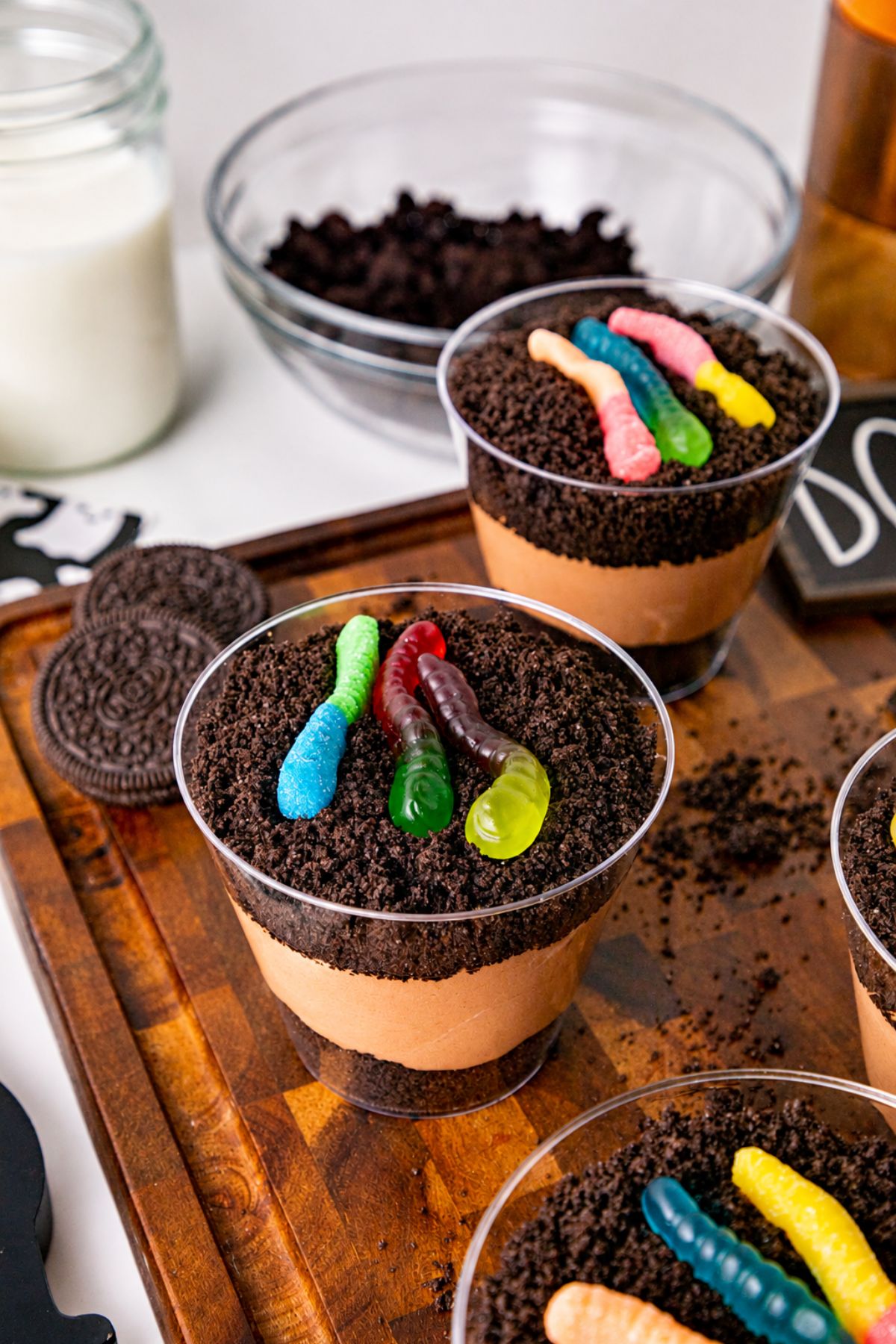 Small individual cups filled with crushed cookies, whipped cream, and chocolate pudding, topped with gummy worms, on a wooden cutting board.