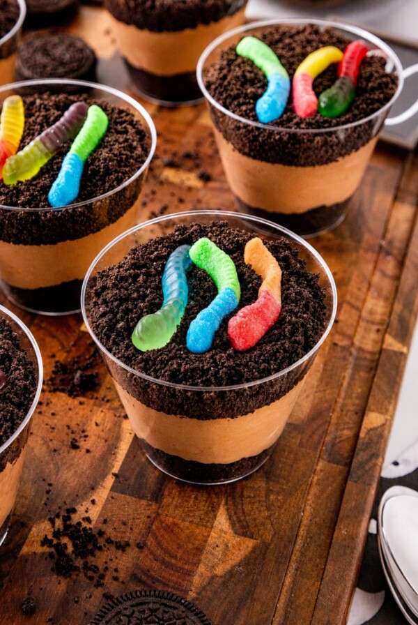 Small individual cups filled with crushed cookies, whipped cream, and chocolate pudding, topped with gummy worms, on a wooden cutting board.