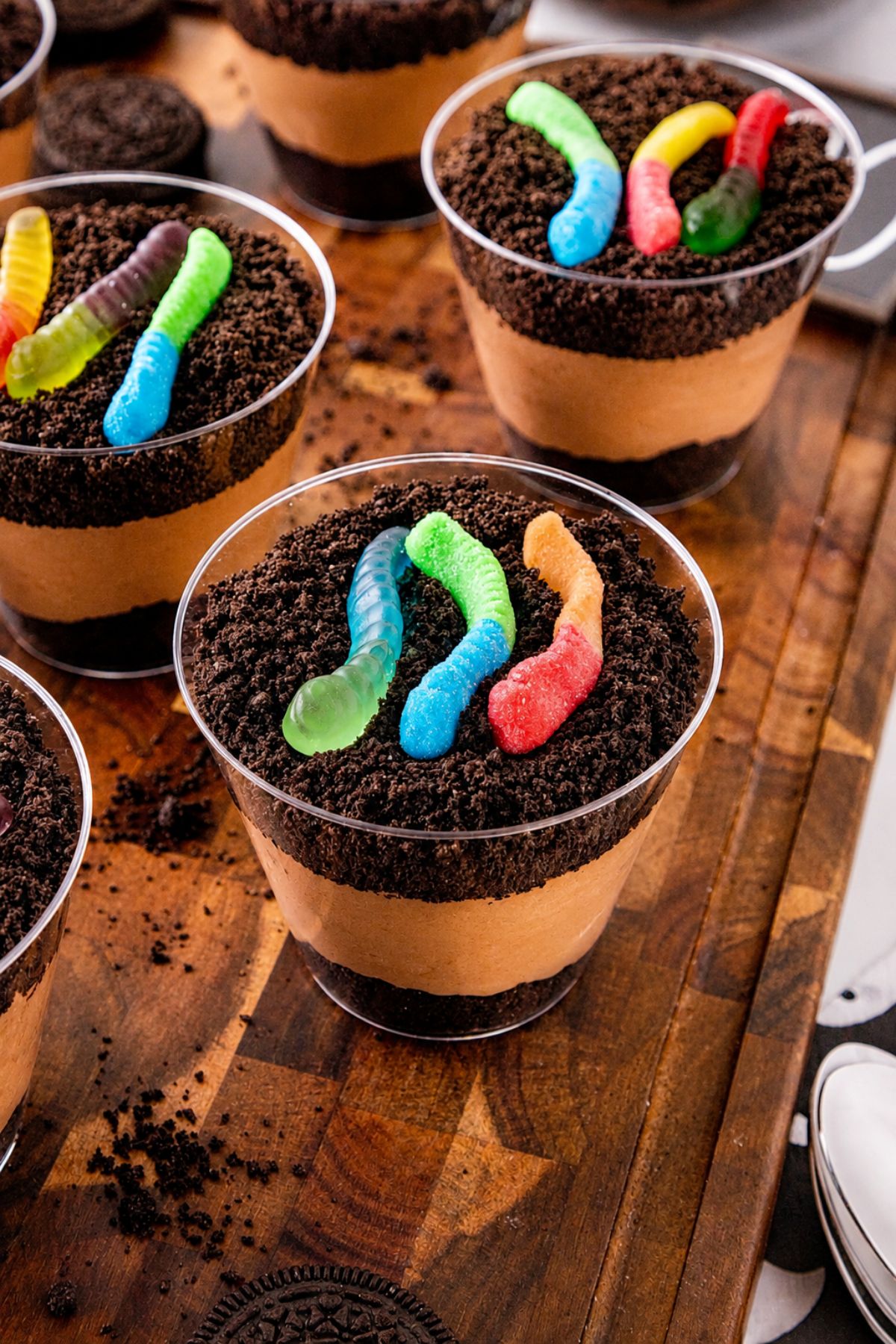 Small individual cups filled with crushed cookies, whipped cream, and chocolate pudding, topped with gummy worms, on a wooden cutting board.