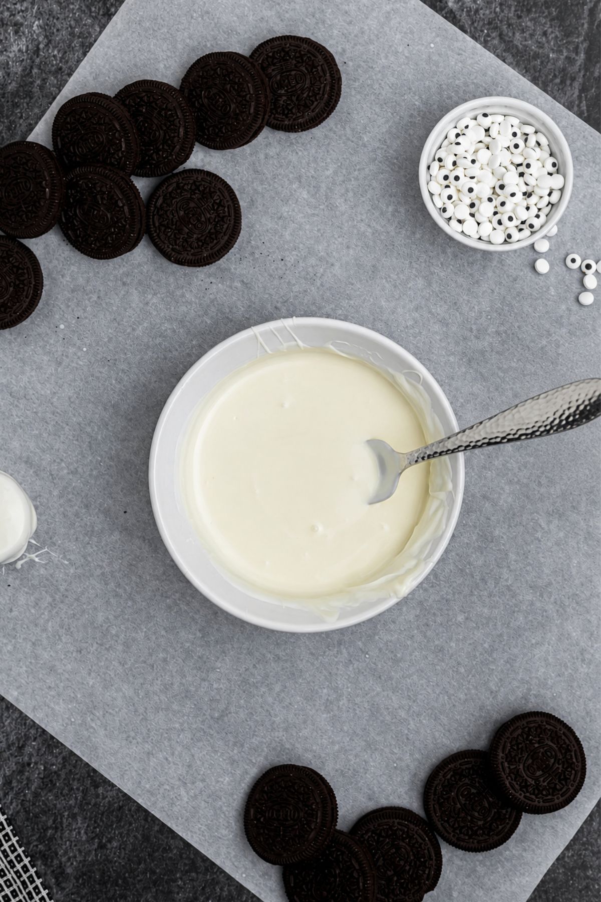 White chocolate melted in a small, shallow bowl, with Oreo cookies on the table, on parchment paper. 