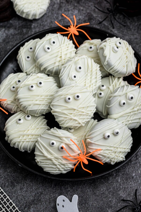 Oreo cookies, dipped and drizzled in white chocolate, with candy eyes, on a black plate, with orange spiders as decoration, on a black marble table.