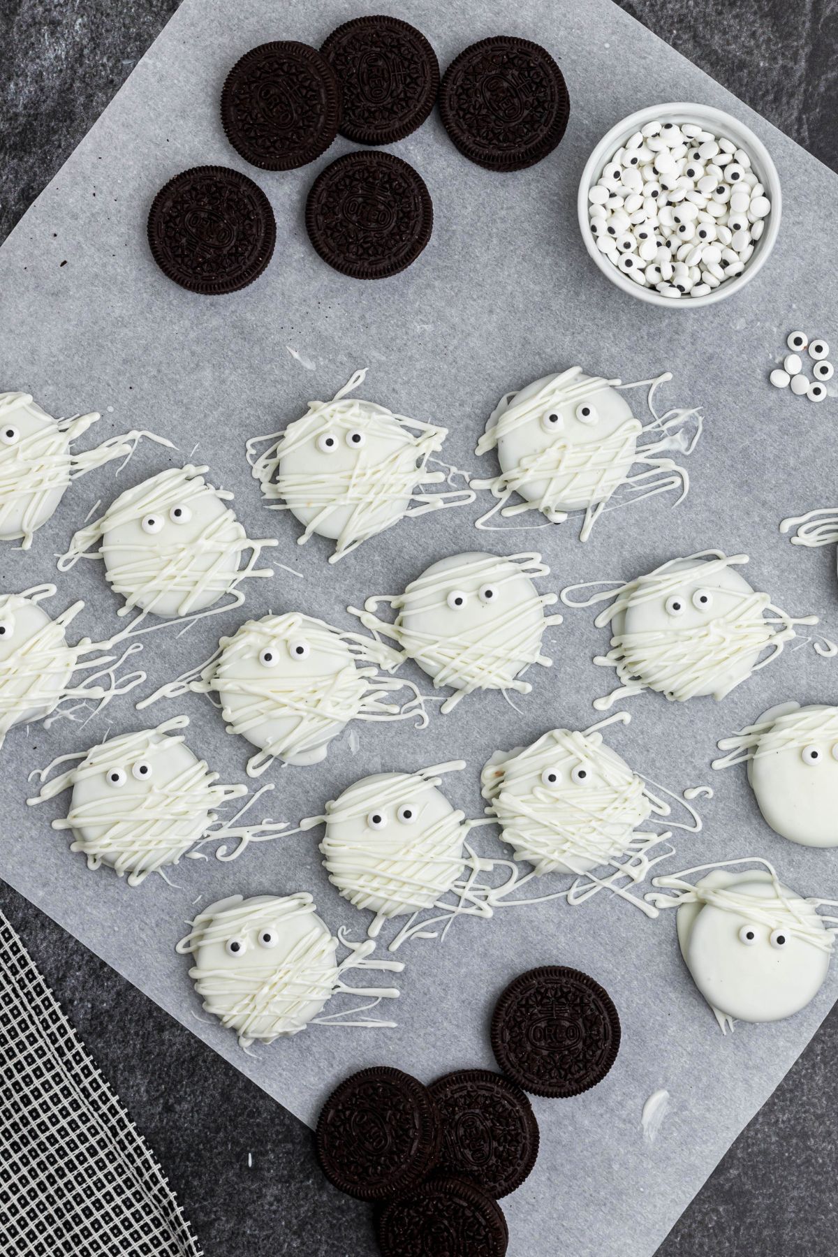 Oreos dipped in white chocolate, drying on parchment paper, with Oreos scattered on the table, with candy eyes added. 