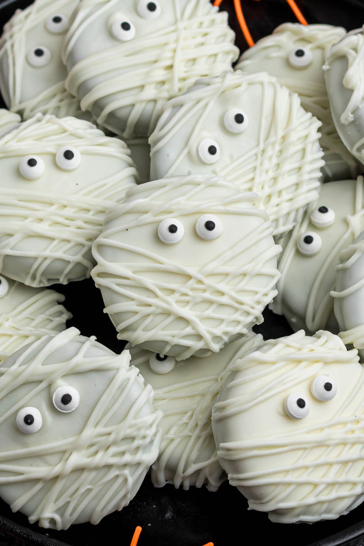 Oreo cookies, dipped and drizzled in white chocolate, with candy eyes, on a black plate, with orange spiders as decoration, on a black marble table.
