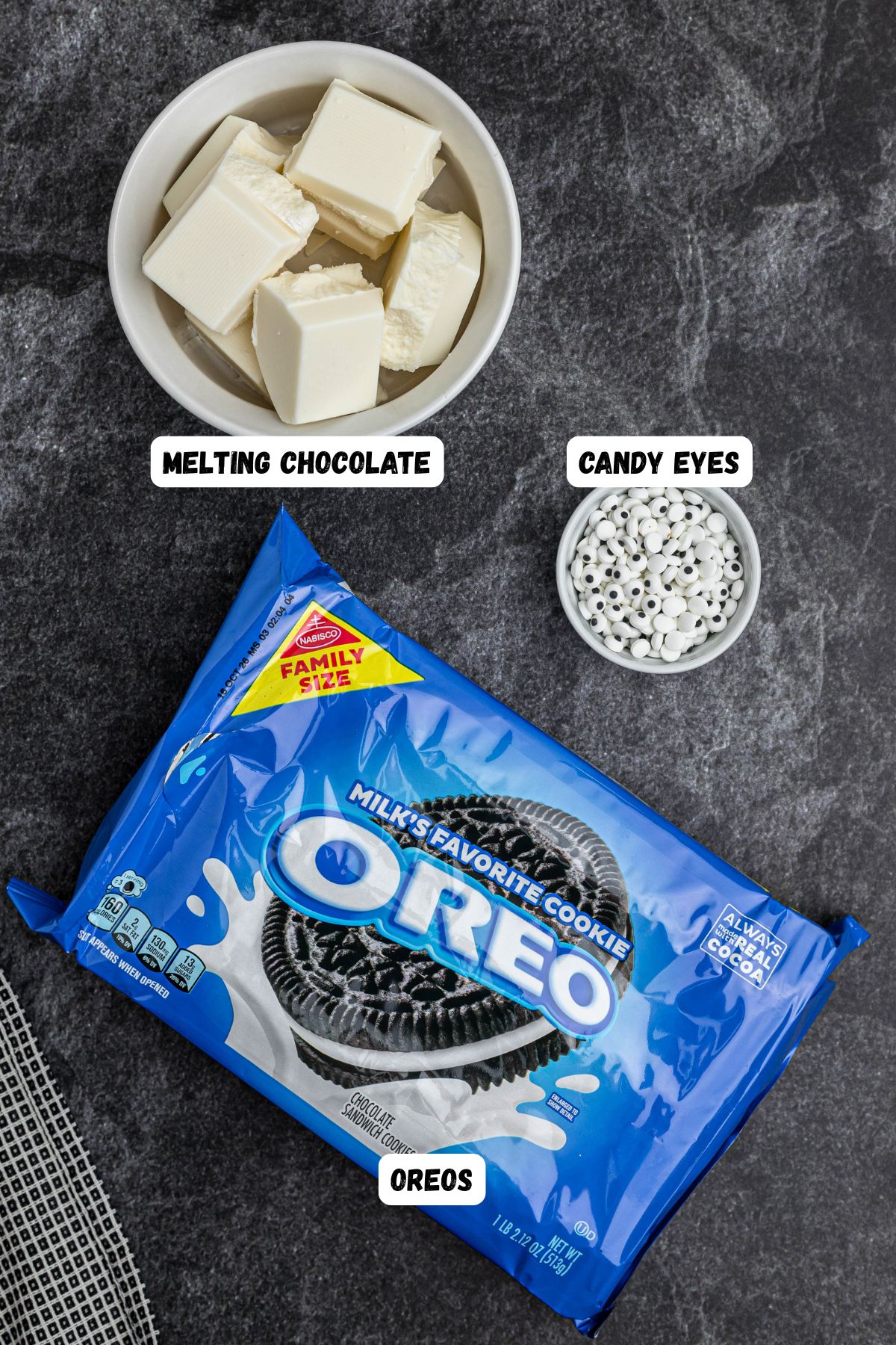 Oreo Mummy Cookies ingredients of Oreos, white chocolate, and candy eyes, on a black marble table, labeled. 