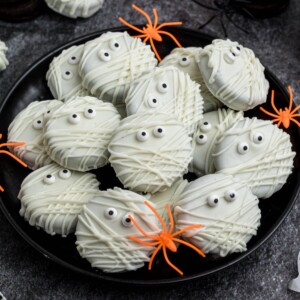 Oreo cookies, dipped and drizzled in white chocolate, with candy eyes, on a black plate, with orange spiders as decoration, on a black marble table.