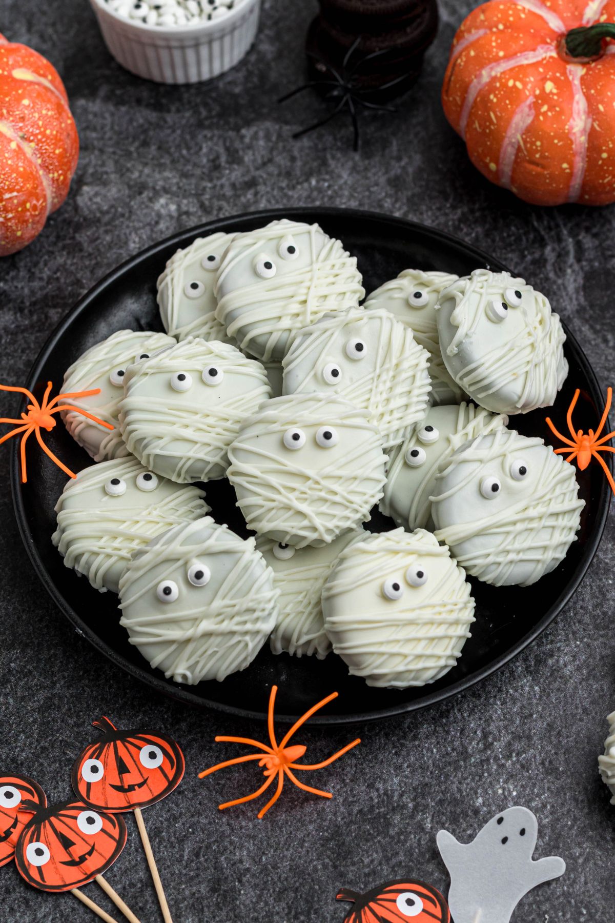 Oreo cookies, dipped and drizzled in white chocolate, with candy eyes, on a black plate, with orange spiders as decoration, on a black marble table. 