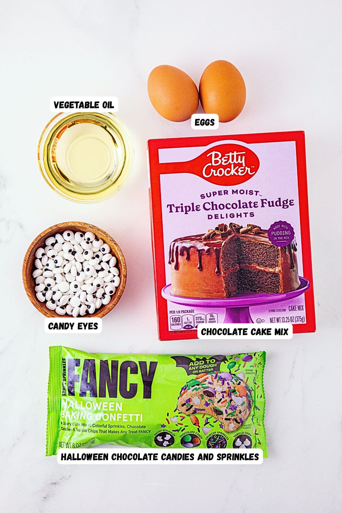 Ingredients needed to make monster cake mix cookies on a white marble table, labeled.