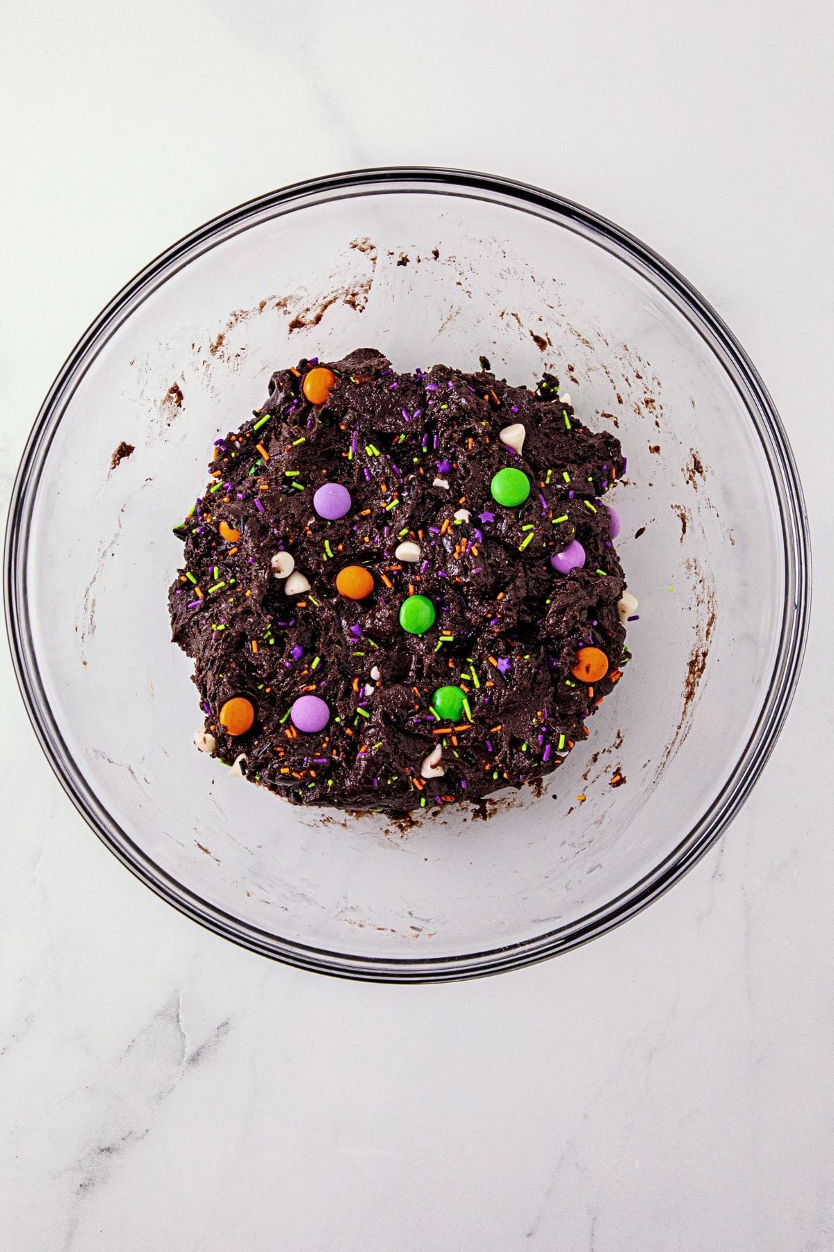 Chocolate cake mix cookie batter, in a large clear glass bowl, with Halloween sprinkles and candies mixed in.
