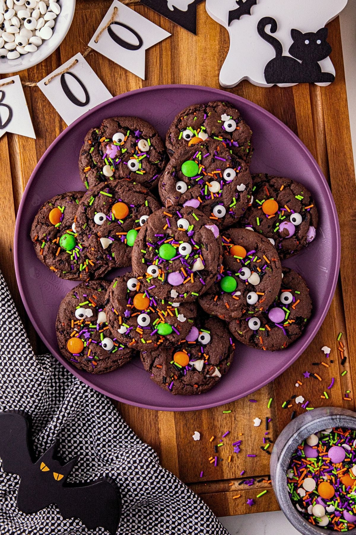 Purple plate with chocolate cookies filled with Halloween sprinkles and candy eye balls, on a wooden cutting board.