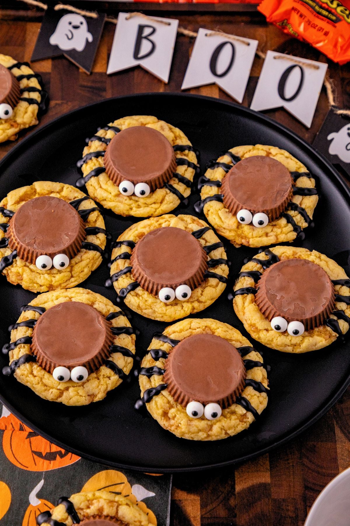 Black plate with spider cookies stacked around the plate, on a table with Halloween decorations scattered around the plate. 