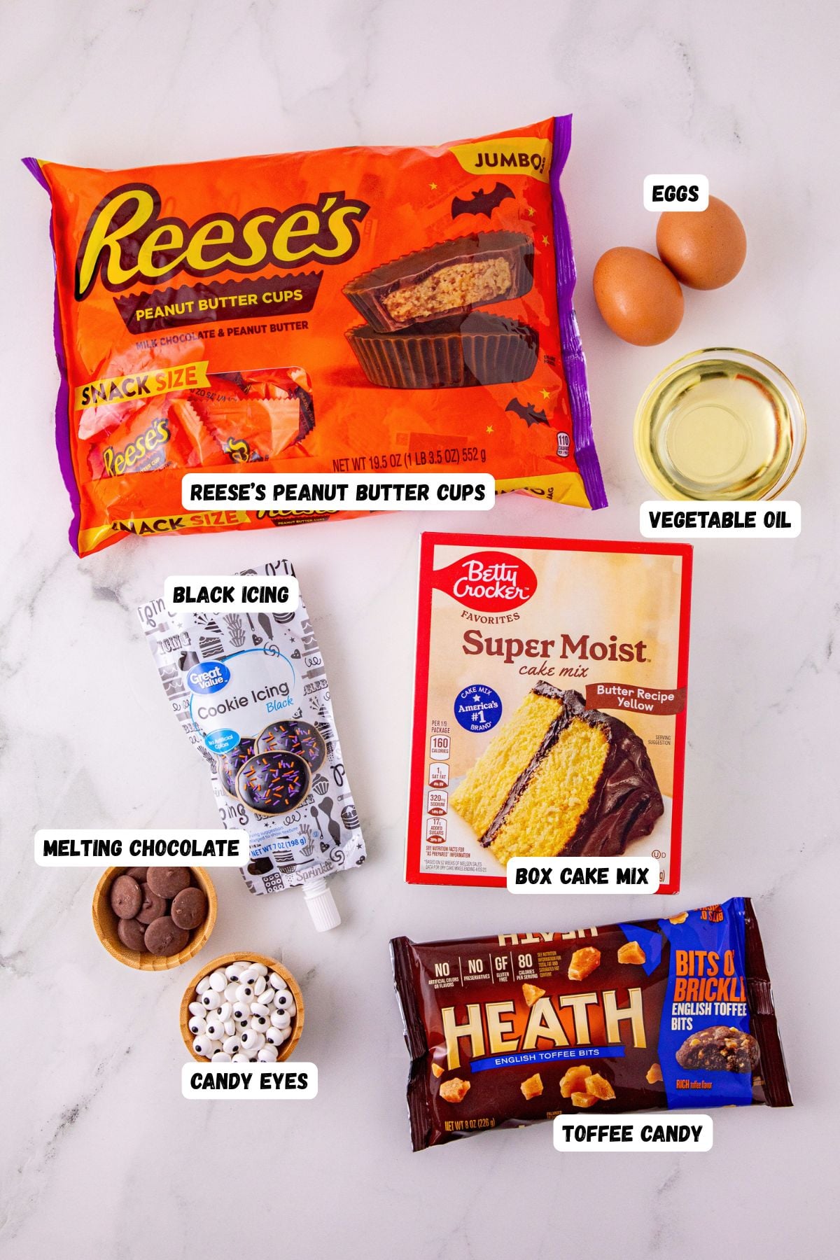 Ingredients like cake mix, eggs oil, black icing, and candy eyes, on a marble table, labeled. 
