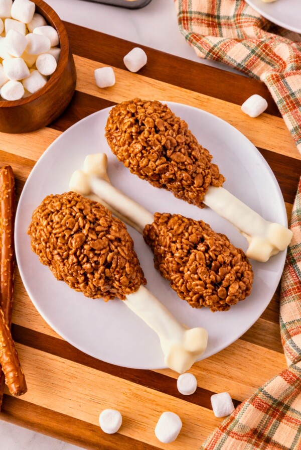 Cocoa krispie treats shaped around white chocolate dipped pretzel rods and marshmallow tips to look like turkey drumsticks. On a white plate, set on wooden cutting board, with marshmallows scattered around.