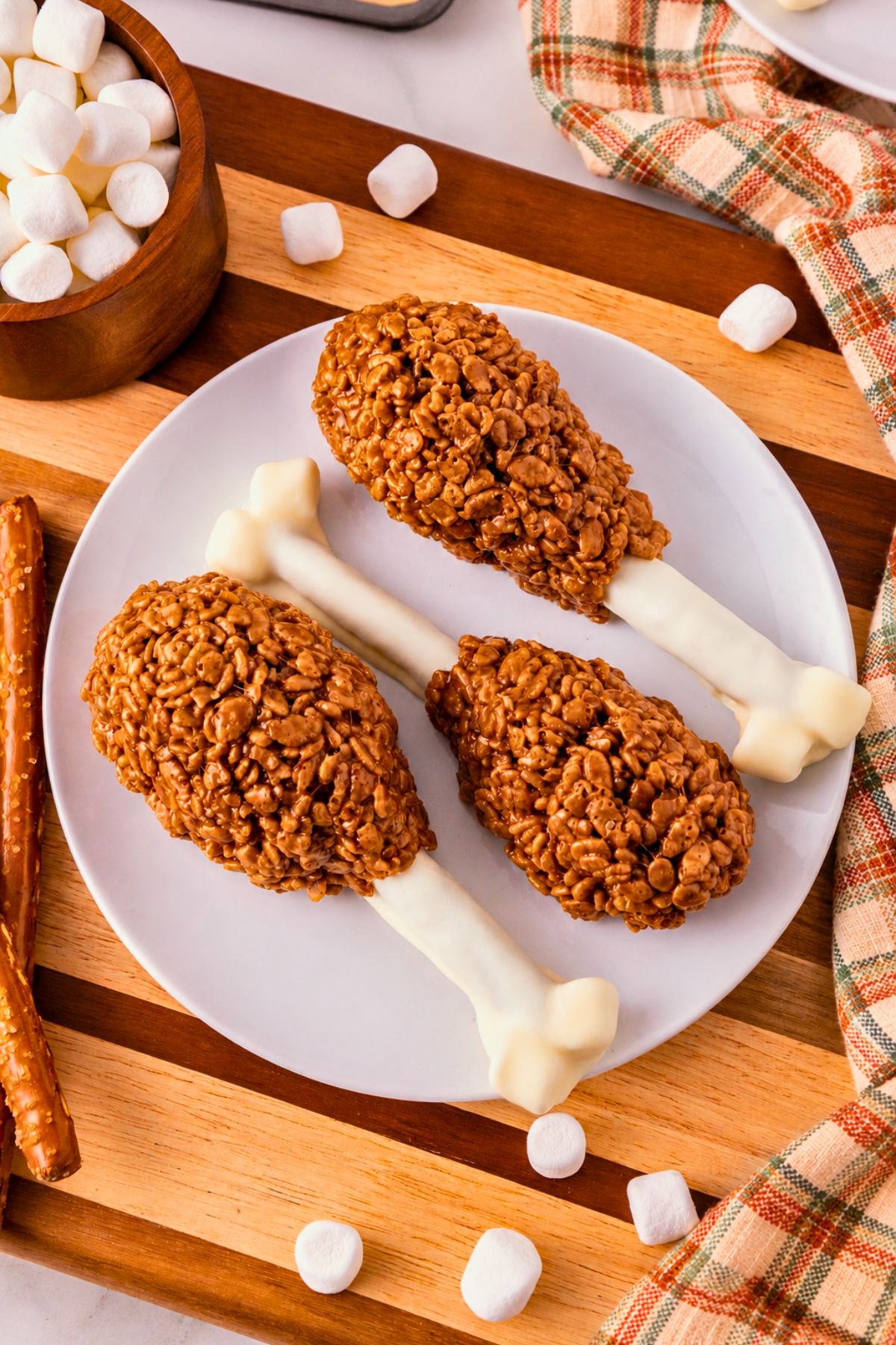 Cocoa krispie treats shaped around white chocolate dipped pretzel rods and marshmallow tips to look like turkey drumsticks. On a white plate, set on wooden cutting board, with marshmallows scattered around. 