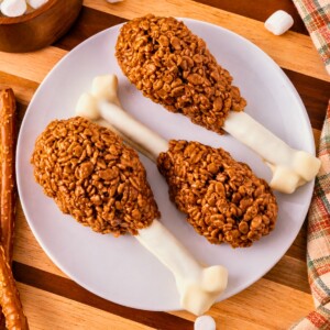Cocoa krispie treats shaped around white chocolate dipped pretzel rods and marshmallow tips to look like turkey drumsticks. On a white plate, set on wooden cutting board, with marshmallows scattered around.